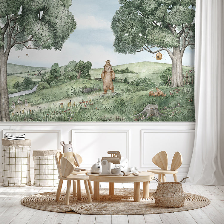 A cozy room adorned with ‘Bears and Bees, Animal Mural Wallpaper’. The room features natural furniture including a round wooden table, chairs, and wicker baskets, amidst a lush landscape on the wallpaper.