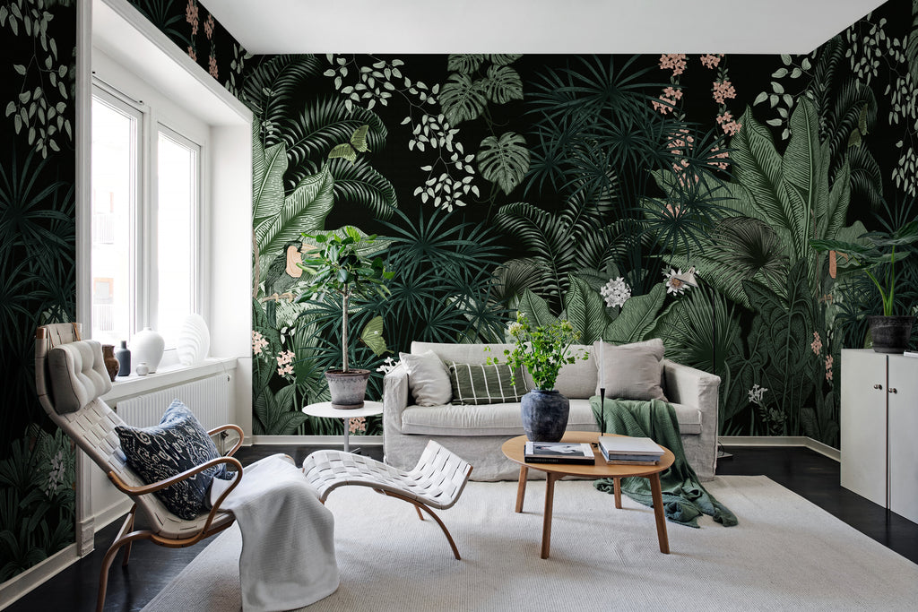 Big Furada, Tropical Mural Wallpaper in black, featured on a wall of a living room with white sofa that has multiple pillows, a wooden round table that has a marble vase with books, and a white relaxing chair