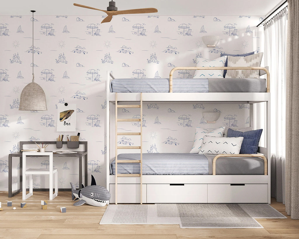 A children’s bedroom exudes tranquility with a white bunk bed adorned with cozy blue and white bedding. The room is illuminated by a woven pendant light hanging from the ceiling, and a ceiling fan ensures a comfortable atmosphere. A plush shark toy adds a playful touch to the room, enhancing its charm. The room’s decor is complemented by a Coastal Beach Day, Pattern Wallpaper Blue, subtly introducing a nautical theme.