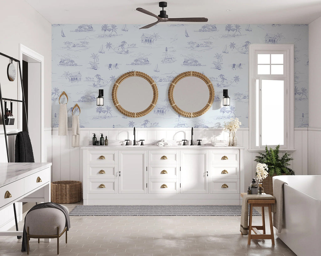 A coastal-themed bathroom with a double vanity and nautical mirrors, lit by a window and a lantern-style light. Coastal Shorelines, Pattern Wallpaper in Blue adds a serene maritime ambiance.