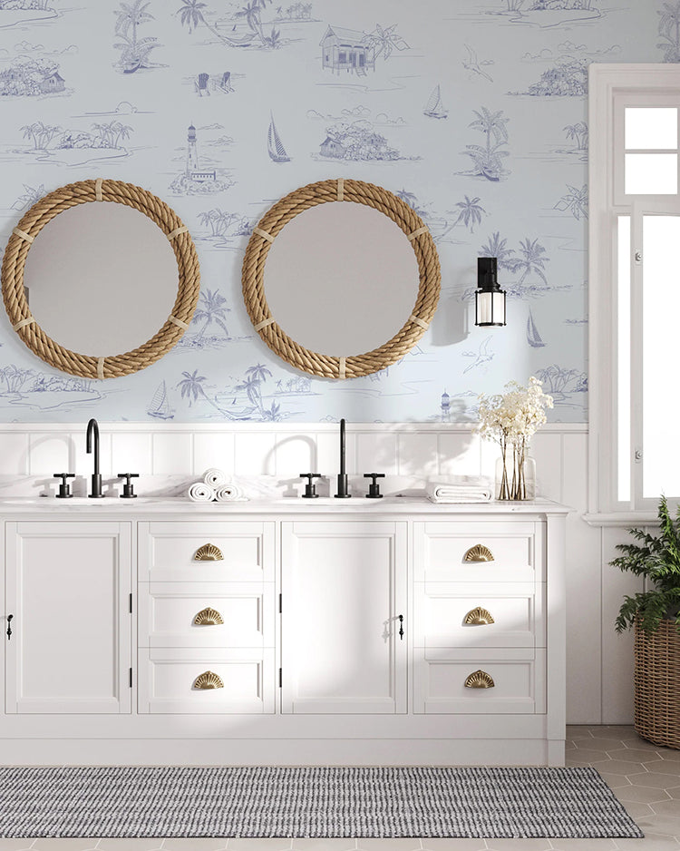 A coastal-themed bathroom with a double vanity and nautical mirrors, lit by a window and a lantern-style light. Coastal Shorelines, Pattern Wallpaper in Blue adds a serene maritime ambiance.