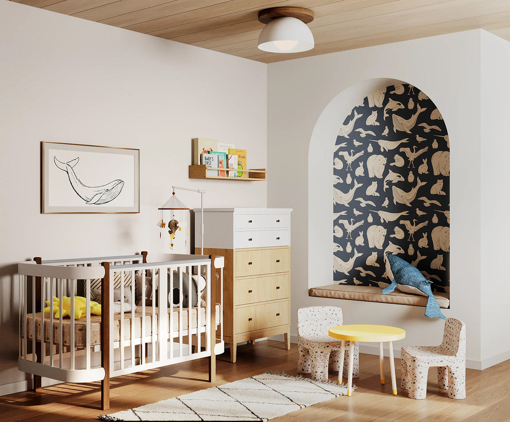 A modern nursery with wooden flooring, a white crib, matching dresser, and shelves filled with toys and books. A whimsical whale illustration adorns the wall. The room is accented by a round yellow table and two small chairs.. The niche features the Frosty Friends, Animal Pattern Wallpaper in Blue.