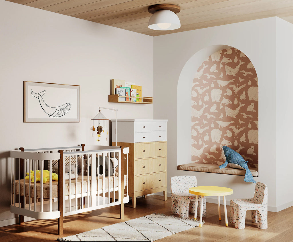 A modern nursery with wooden flooring, a white crib, matching dresser, and shelves filled with toys and books. A whimsical whale illustration adorns the wall. The room is accented by a round yellow table and two small chairs.. The niche features the Frosty Friends, Animal Pattern Wallpaper in dusty pink.