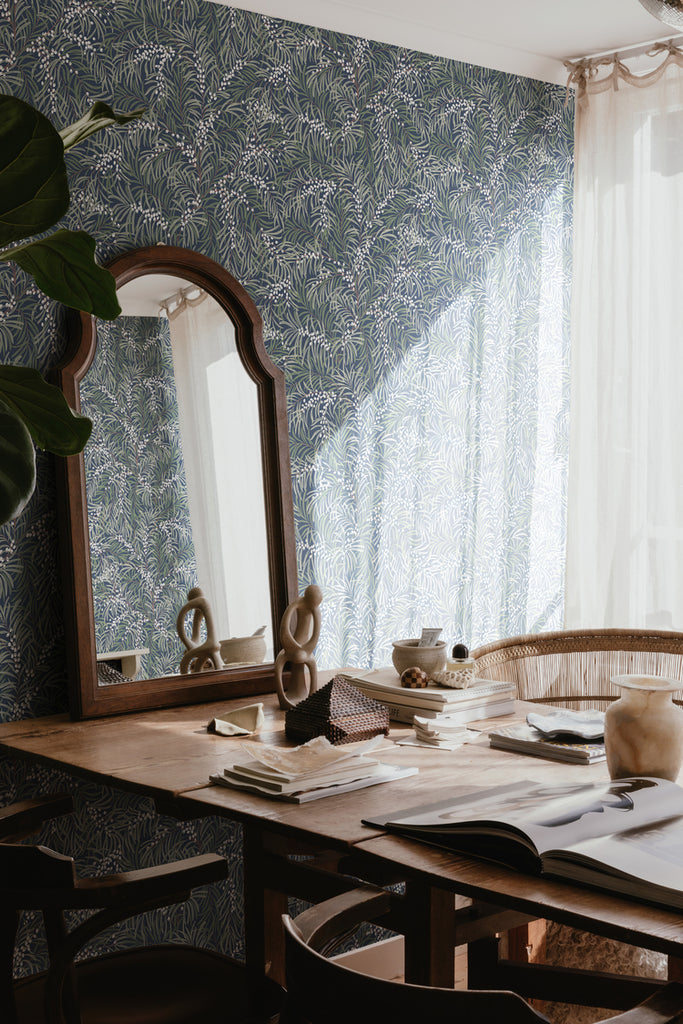 Idun, Nature Pattern Wallpaper in dark blue featured on a wall of a room with wooden table with books, ceramics, and mirror on top, and a Rattan Chair. 