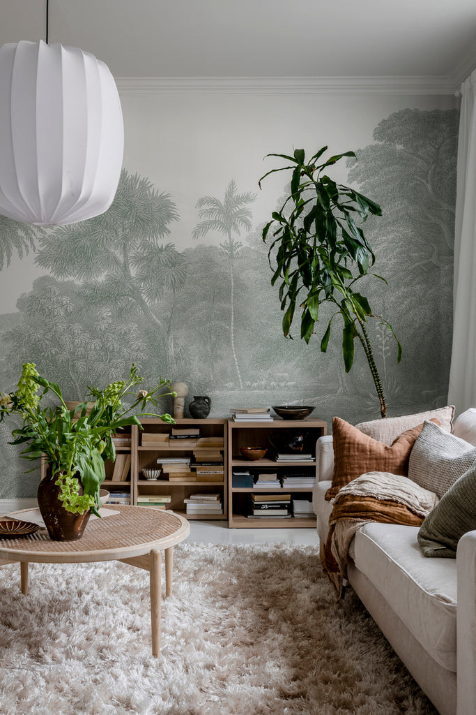 Jungle Land, Mural Wallpaper in fLight Grey featured on a wall of a living room with white sofa, brown round table and cabinet full of books