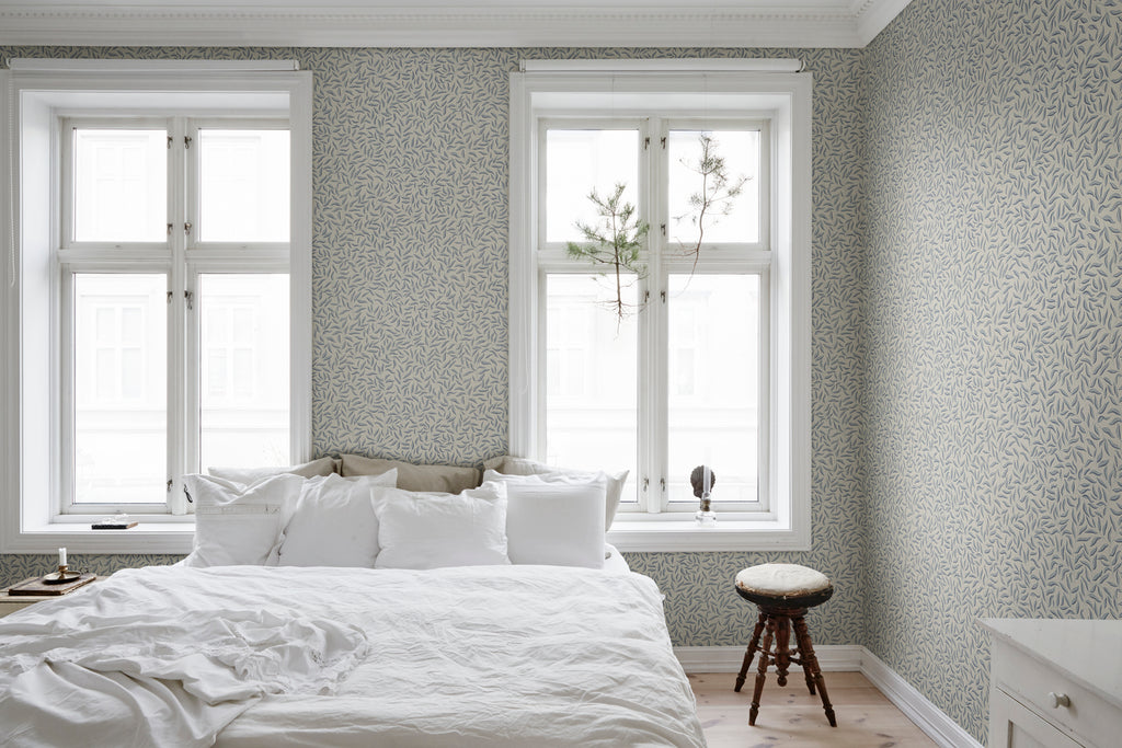 Karolina Foliage, Pattern Wallpaper in dark blue featured on a wall of a bedroom with a clean white bedsheet and pillows and a side stool