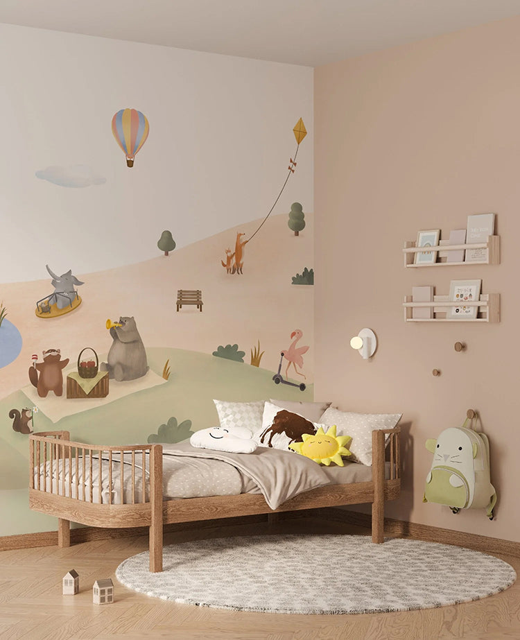 A child’s room with Playground Adventures, Animal Mural Wallpaper on one wall. A wooden crib with plush toys, a small white shelf with books, a grey rug, and a green backpack on the wall