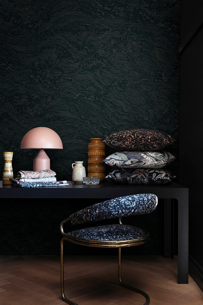 Sea Foam, Wallpaper in Dark Green featured on a wall of a room, with black table that has multiple patterned pillow, and pink lamp, with a patterned chair next to it. 
