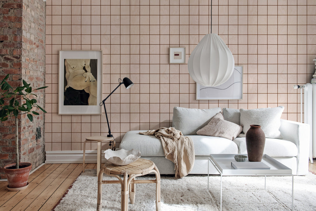 Featuring Squares and Tiles illustrations, a Wallpaper in Nude adorns a living area wall. The space includes a white sofa with multiple pillows on a furry mat, a white table with a brown vase, and a nearby rattan chair