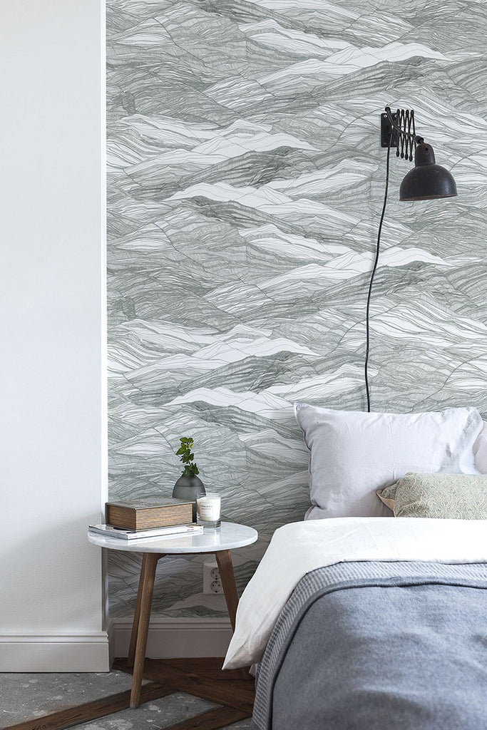 The bedroom features a wall with Tidal Waves Pattern Wallpaper in Grey, a bed dressed in grey sheets with a white pillow, and a side table adorned with a book and a ceramic container.