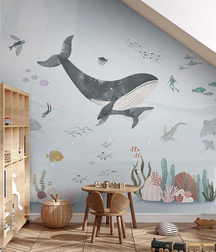 A vibrant children’s room featuring a ‘Whale and Ocean Friends’ mural featuring whales, pufferfish, flying fish, clown fish, stingray, divers and more. The room is filled with a small table and chairs, a variety of toys, and a bookshelf. The wooden floor adds a warm touch to the playful atmosphere.