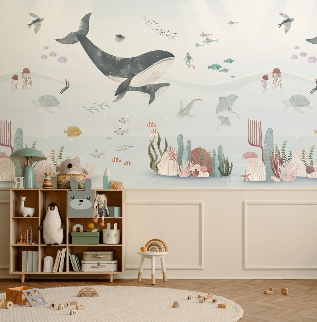 A vibrant children’s room featuring a ‘Whale and Ocean Friends’ mural featuring whales, pufferfish, flying fish, clown fish, stingray, divers and more.  The room features a polished wooden floor, crisp white wainscoting, and a neatly arranged shelf filled with toys, creating a warm and inviting atmosphere.
