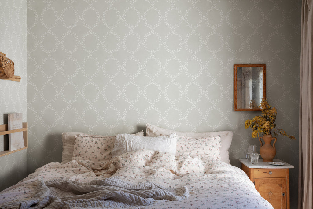 Wilma Wreath Patterned Wallpaper in Grey Featured on a wall of a bedroom with patterned bedsheets and pillows with a wooden side table that has a flower vase