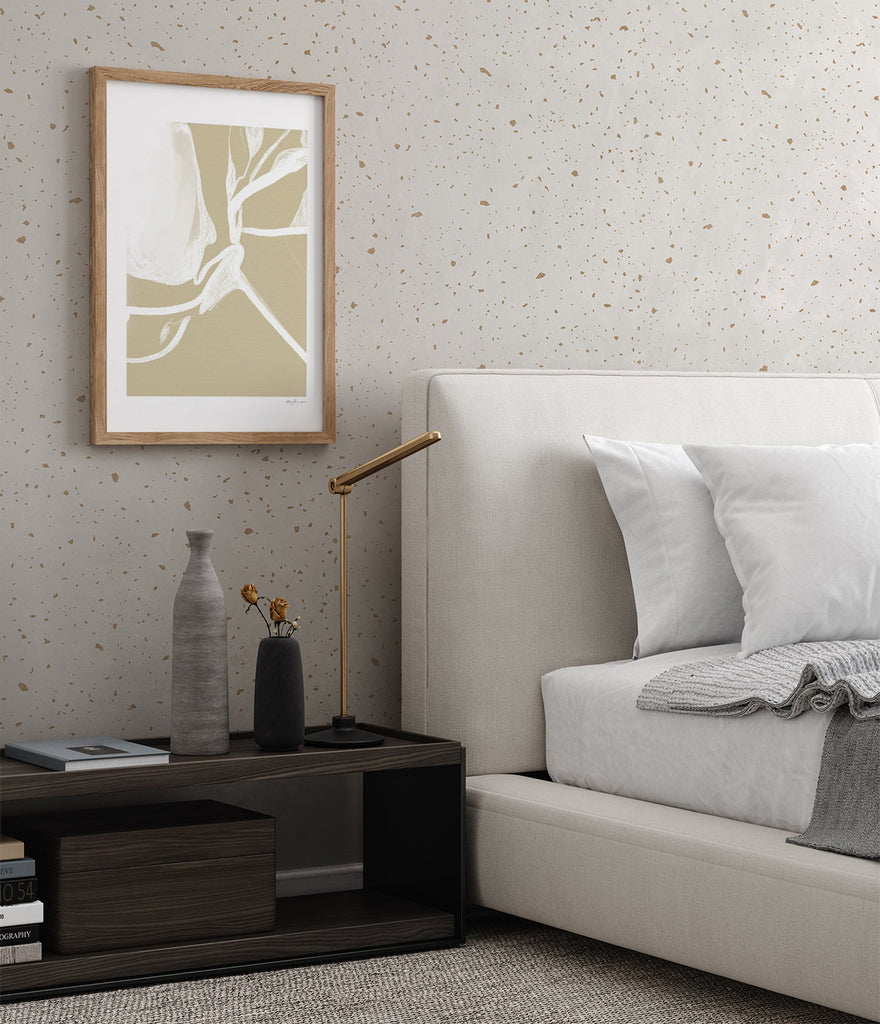 Gold metallic confetti speckles wallpaper applied in a bedroom with a dark wood side table and a grey bed.