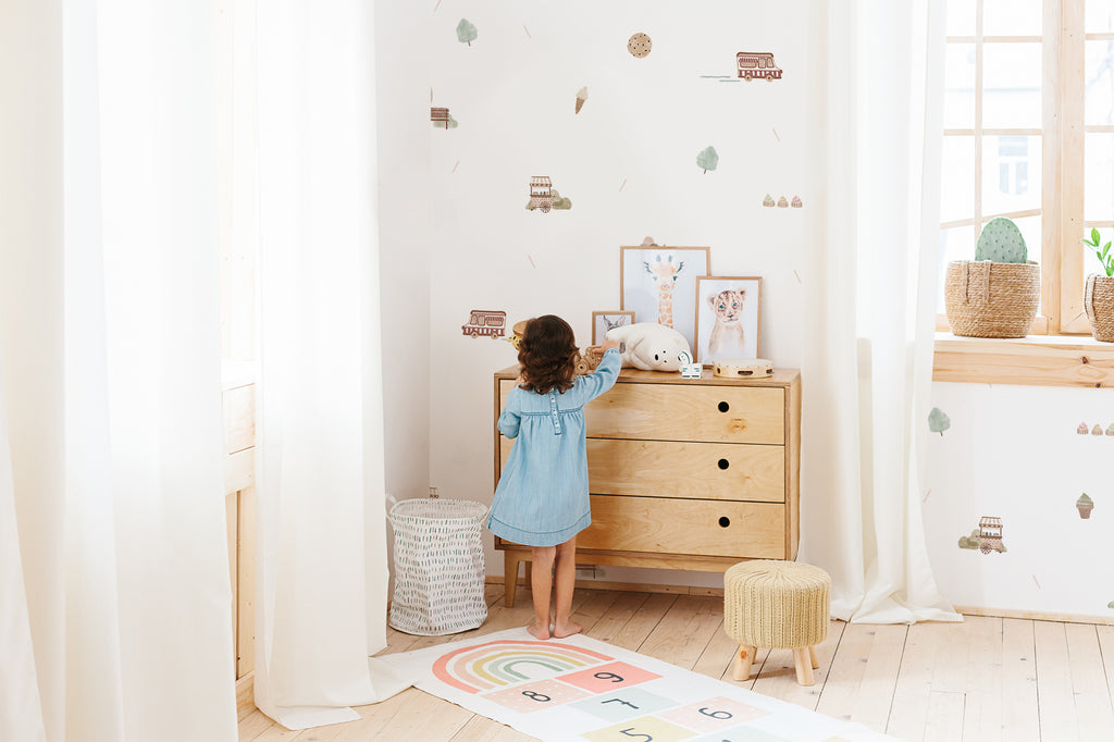 Brightly lit playroom with a little girl in a blue dress, with light coloured furnishings. On the wall is a white wallpaper with sweet treats motifs. 
