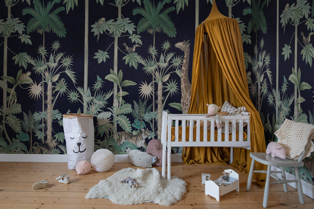 A Walk in the Jungle, Mural Wallpaper in dark blue as seen on a wall of a child’s playroom with white crib and mustard yellow fabric 