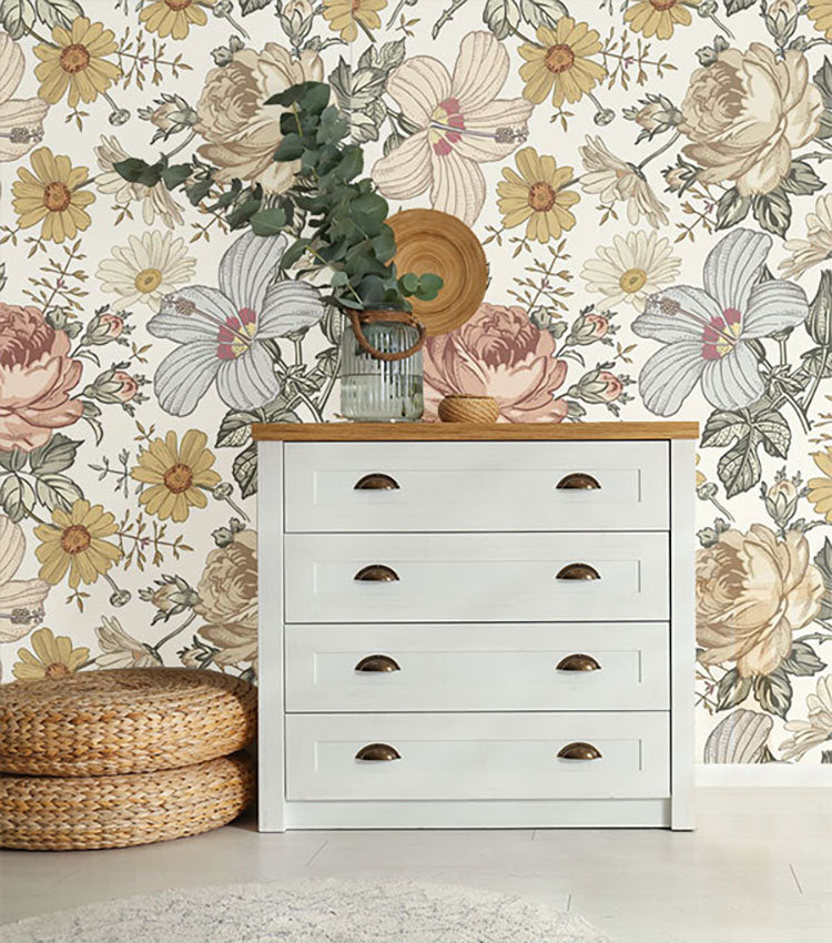 Adora, Vintage Floral Mural Wallpaper in Sand adorning a room, featuring large, hand-drawn blooms in hues of pink, yellow, and white. A white dresser with round handles and a woven basket with greenery on top enhance the room’s aesthetic.