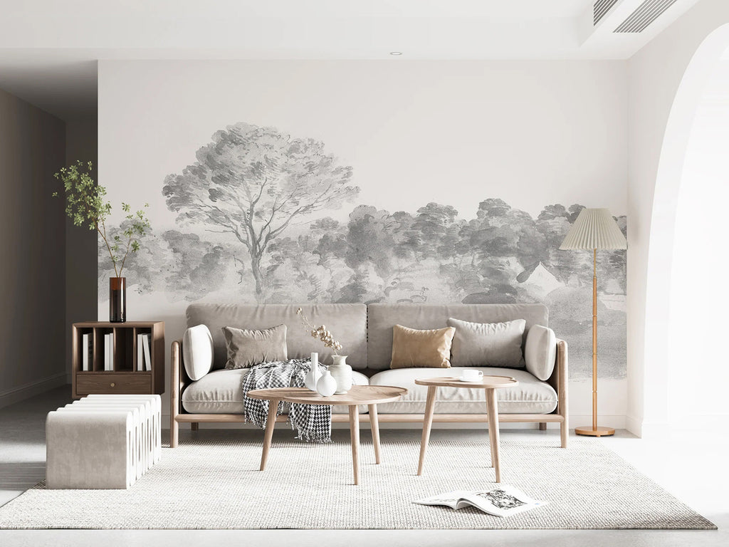 A cozy living room bathed in soft light, adorned with An Old World Arboretum watercolour mural in black and white wallpaper. The room is furnished with a plush beige sofa laden with cushions, a rustic wooden coffee table, and a side table stacked with books. A floor lamp casts a warm glow, enhancing the room’s inviting ambiance.