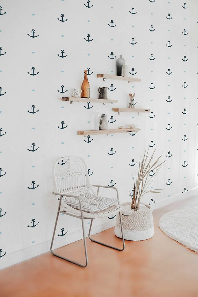 A nautical-themed room, adorned with a modern white chair and wooden shelves displaying an array of decorative items. A woven basket with tall dried grasses adds a rustic touch. The room’s charm is enhanced by the Anchor Pattern Wallpaper, adding a maritime feel.
