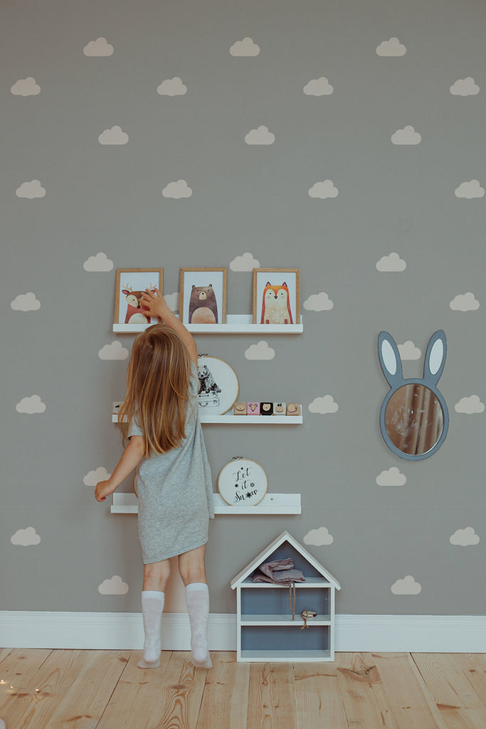 A child in a gray dress reaches for picture frames on a shelf against a wall adorned with Asaph Clouds pattern wallpaper, featuring fluffy white clouds on a soft gray background.
