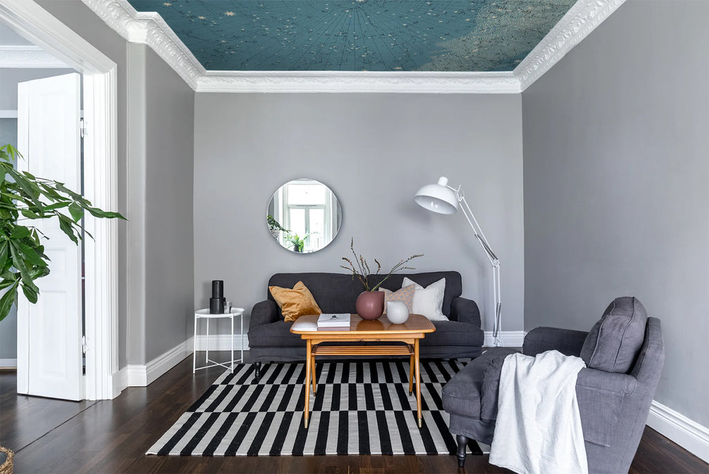 An elegantly decorated living room with a dark grey sofa, matching armchair, and wooden coffee table on a striped rug. The ceiling showcases the captivating ‘Atlas of Astronomy’ star map mural wallpaper, adding celestial charm to the space.