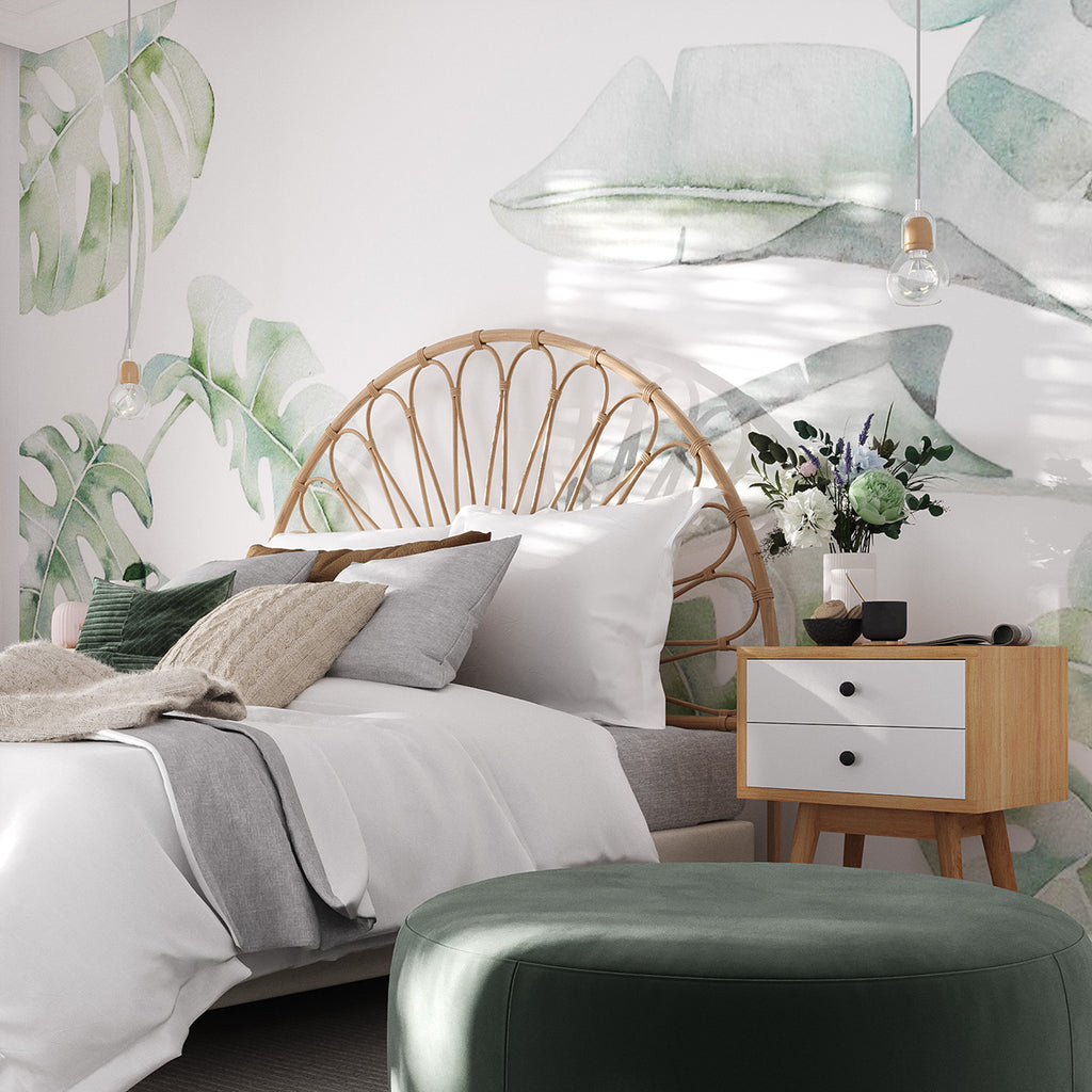 A serene bedroom featuring the Garden Bloom, Tropical Mural Wallpaper with a botanical design. The room includes a rattan headboard, white bedding, green pillows, a wooden nightstand with flowers, and an emerald green ottoman.