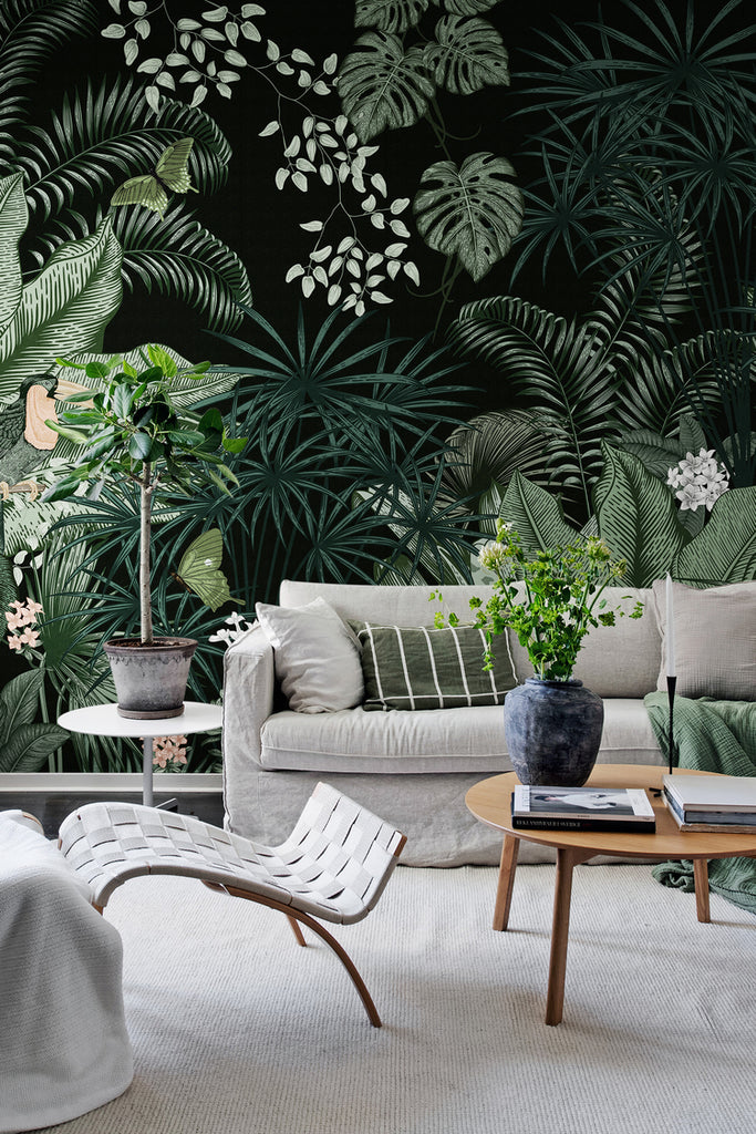 Big Furada, Tropical Mural Wallpaper in black, featured on a wall of a living room with white sofa that has multiple pillows, a wooden round table that has a marble vase with books, and a white relaxing chair