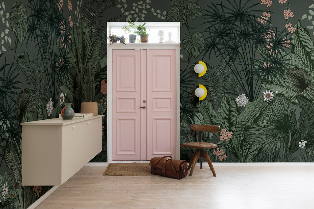 Big Furada, Tropical Mural Wallpaper in green featured on a wall of a room with a pink door, in the room is a suspended cabinet with ceramics and book on top. A wooden stool and a bag can also be seen, on a wood flooring. 