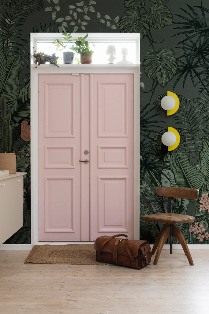 Big Furada, Tropical Mural Wallpaper in green featured on a wall of a room with a pink door, in the room is a suspended cabinet with ceramics and book on top. A wooden stool and a bag can also be seen, on a wood flooring. 