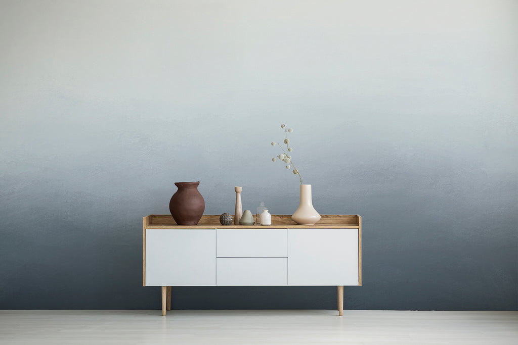 A minimalist room with a wooden console table against a wall featuring the Blake Horizon, Ombre Mural Wallpaper in Blue. Featuring various ceramic vases on a side board.