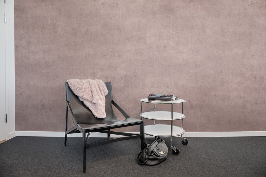 Blush Solid, Concrete Wallpaper featured on a wall of a room with a single chair and table with a black carpet covering the flooring