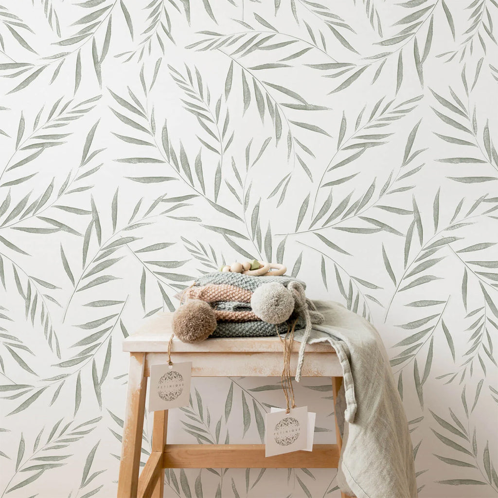 A serene scene with a white wall adorned with Botanical Foliage, Pattern Wallpaper. A wooden stool with cozy, neutral-toned knitwear adds a touch of warmth.