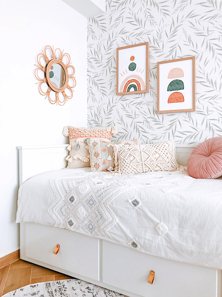 A cozy, well-lit bedroom corner adorned with a Botanical Foliage, Pattern Wallpaper. An rattan oval mirror and two framed artworks hang on the wall. A white bed with various pillows and a round pink cushion is positioned against the wall.