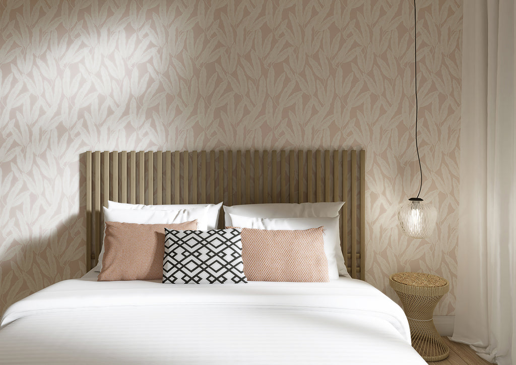 Botanist, Tropical Pattern Wallpaper in Nude featured on wall of a cozy bedroom with a bed that has white bedsheet, and a variety of pillows. It has a wooden headboard and a rattan side table is also visible