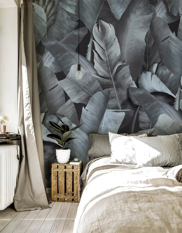 Botany Mist, Tropical Mural Wallpaper featured in a wall of a bedroom with white pillows and sheets, which is adjacent to a window giving the room natural lighting and cozy ambiance. 