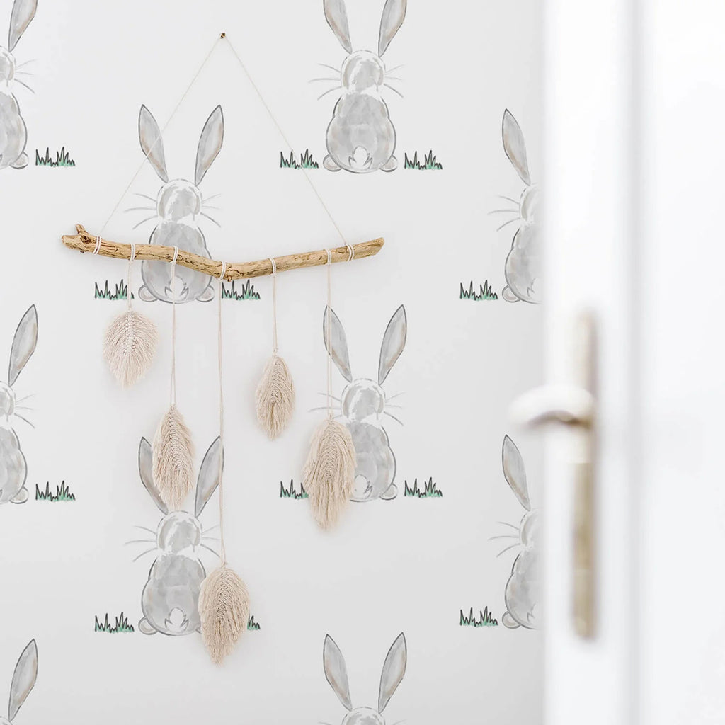 A whimsical wallpaper featuring a Bunny Butt, Pattern Wallpaper on a white backdrop. A boho-style feather wall hanging adds a touch of elegance to the scene. Part of a modern white door or window is visible on the right.