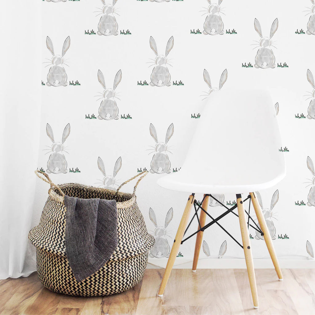 A playful room featuring Bunny Butt, Pattern Wallpaper on a white backdrop. A modern white chair and a woven basket with a grey blanket are in front of the wallpaper. The floor is light-colored wood.