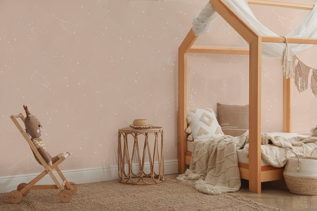 A charming children’s bedroom featuring a wooden canopy bed with white draping. A matching wooden rocking horse adds a playful touch. A woven side table and a textured rug bring warmth and texture to the space. The room is wallpapered in Chalky Stars, Pastel Pattern Wallpaper in Blush Pink, adding a whimsical charm to the serene ambiance.