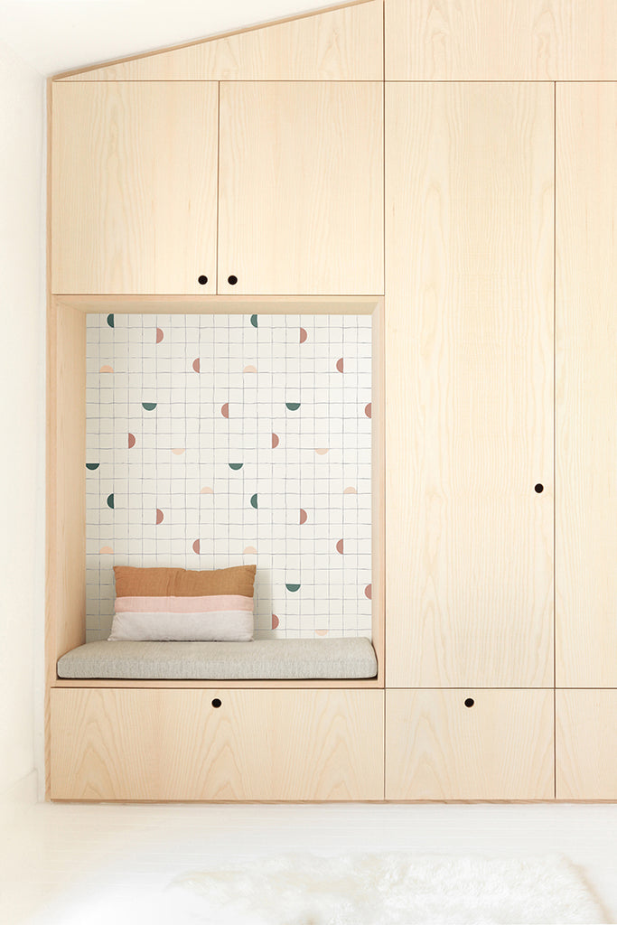 Checkerboard, Geometric Wallpaper in Green/BlushPink/LightPink adorn a cozy nook with a seat cushion, surrounded by wooden panels. The inviting space features a comfortable grey seat cushion and a single rectangular orange pillow. The surrounding light-colored wooden wall unit suggests storage spaces with black circular handles on each door panel.