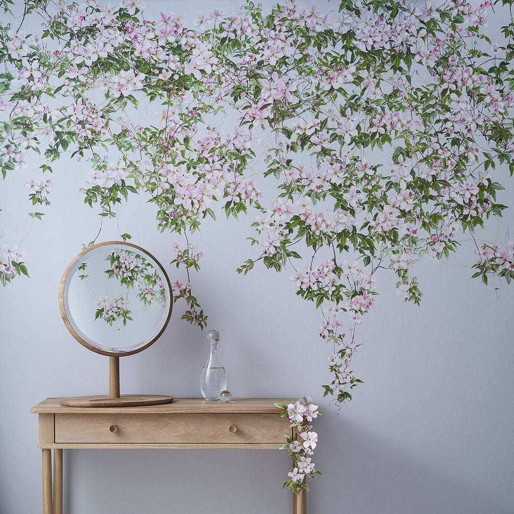 The wall is adorned with Classic Clematis, Floral Mural Wallpaper in Blue that creates a serene and elegant atmosphere. A wooden table holds a round mirror and a clear glass bottle containing a few of the clematis flowers. The mirror reflects part of the mural, blending natural and reflected imagery. 
