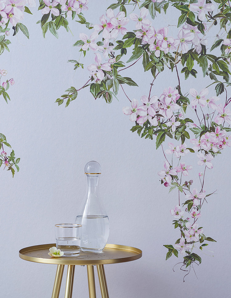 Classic Clematis, Floral Mural Wallpaper in Blue creates an elegant atmosphere. A gold-colored round table holds a clear glass carafe with water and a small white flower. 