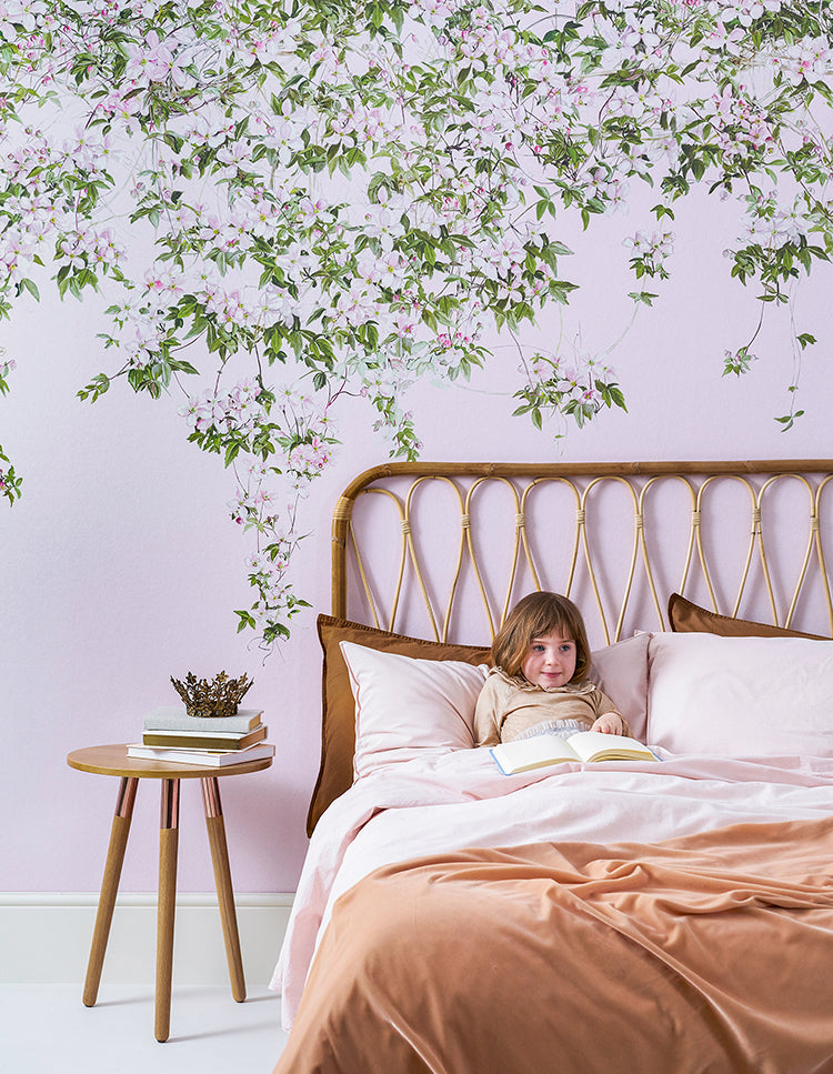 A serene bedroom with a person reading on a bed. The room is adorned with a Cozy bedroom with Classic Clematis, Floral Mural Wallpaper in Pink. A wooden headboard, white sheets, and a brown blanket add to the cozy atmosphere.
