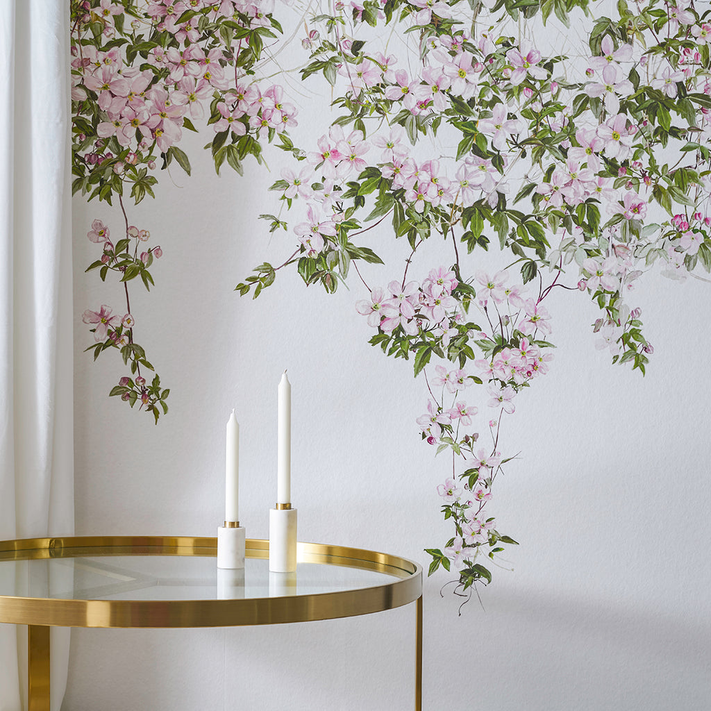 A serene room adorned with Classic Clematis, Floral Mural Wallpaper in White featuring pink flowers and green leaves. A golden round table with two tall white candles in golden holders adds to the elegance. A part of a white curtain suggests a window nearby