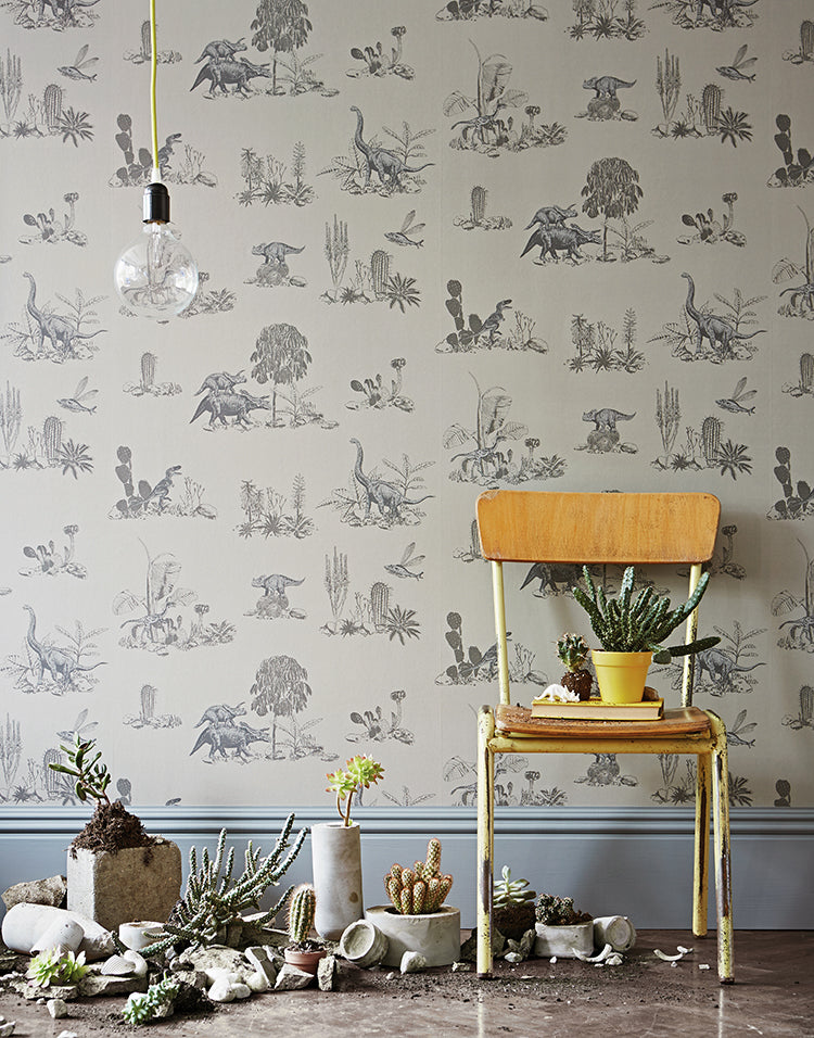 Classic Dino, Pattern Wallpaper in Grey featured on a wall with a vintage chair & potted plants against a wall adorned with intricate dinosaur illustrations, under a hanging bulb.
