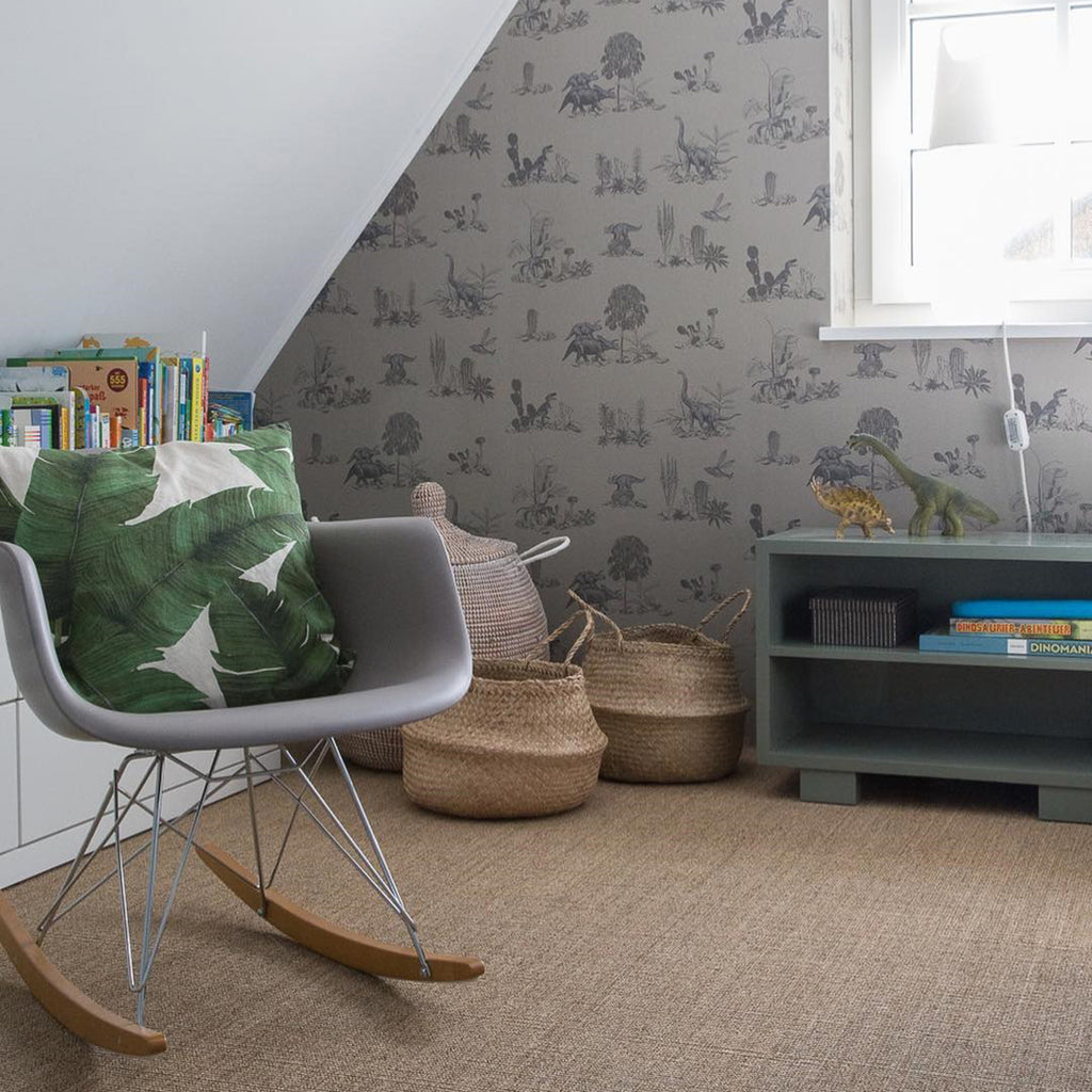Classic Dino, Pattern Wallpaper in Grey featured on a wall bathed in natural light from a window. A modern gray rocking chair with wooden rockers is adorned with a leaf designed cushion. A small dark green bookshelf holds books and an ornamental item, and woven baskets add a homely touch. 