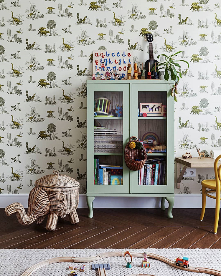 The wall is adorned with a Classic Dino, Pattern Wallpaper in Green, creating a playful backdrop. A mint green cabinet filled with colorful books and toys stands against this wall, with more toys and potted plants on top. In front of the cabinet is a woven basket shaped like a elephant, adding to the room’s playful theme. 