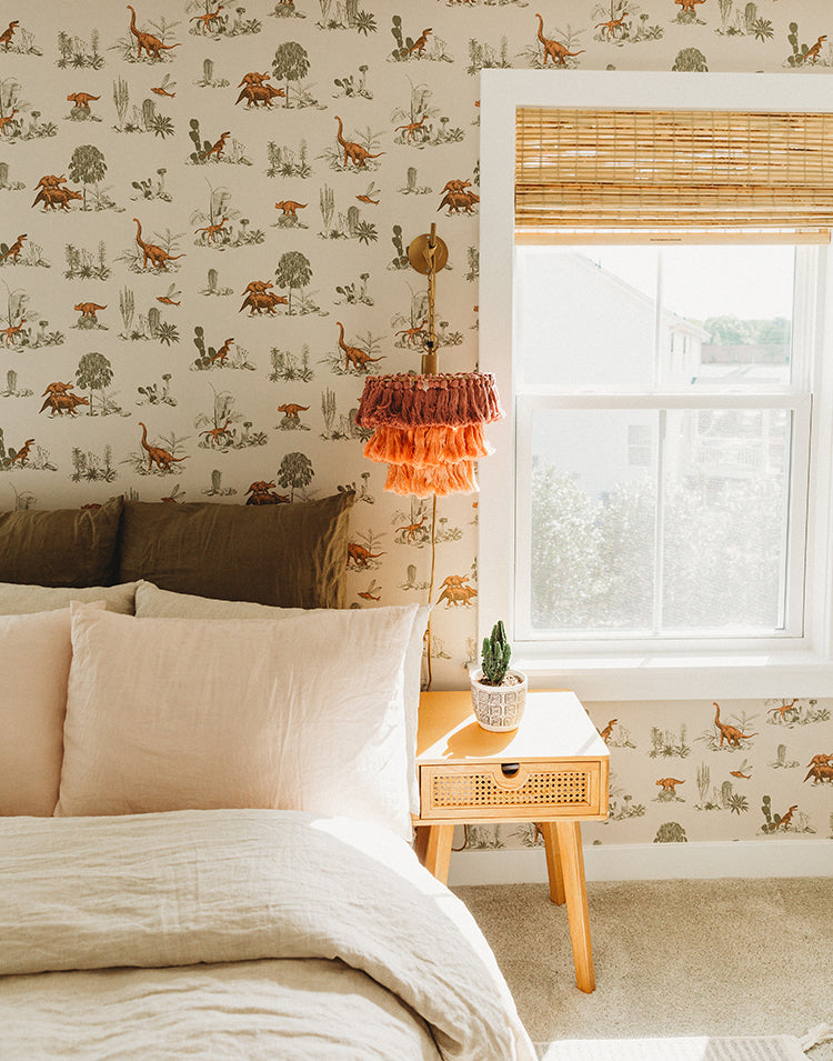 Classic Dino, Pattern Wallpaper in Pink featured on a wall of a cozy bedroom corner with a bed, a window with bamboo blind, and a hanging fringe lamp. A small wooden table holds an ornamental plant.