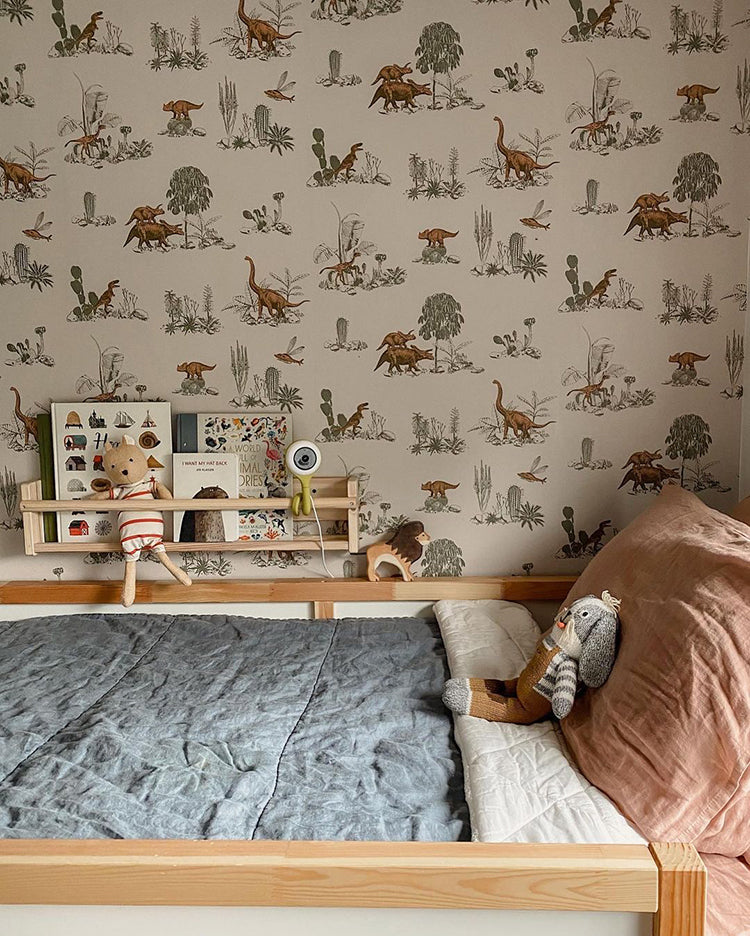 Classic Dino, Pattern Wallpaper in Pink featured on a wall of a cozy bedroom setting with a neatly made bed covered with a blue quilt and terracotta pillows. A plush toy sits on the bed. To the left of the bed, there’s a small wooden shelf filled with various toys and decorative items. 