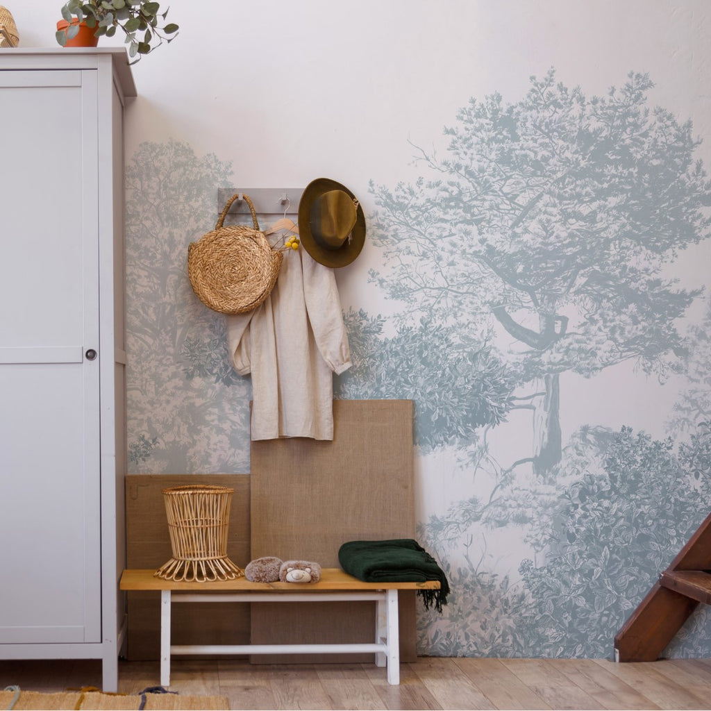 Classic Mua, Mural Wallpaper in Blue creates a cozy corner with a wooden bench, woven basket seat, coat rack with a straw bag, hat, and clothing, and a grey door or cabinet with potted plants.”
