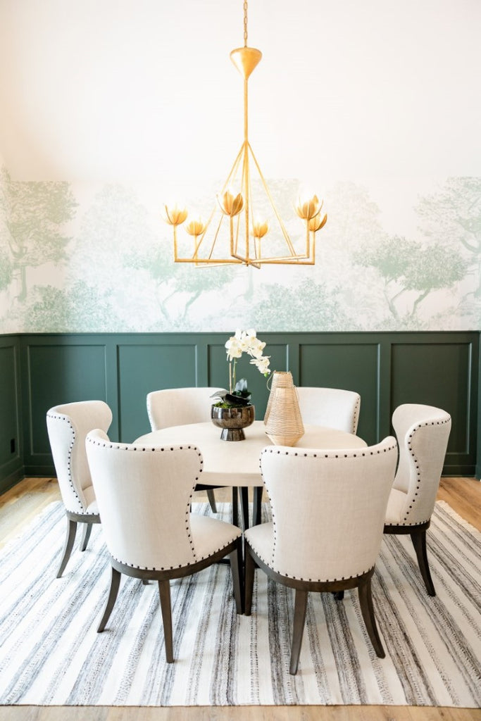 Classic Mua, Mural Wallpaper in Green brings a nature-inspired aesthetic to a well-lit dining area with a golden chandelier, a round wooden dining table surrounded by white upholstered chairs, a vase with white flowers, and a striped rug on the floor
